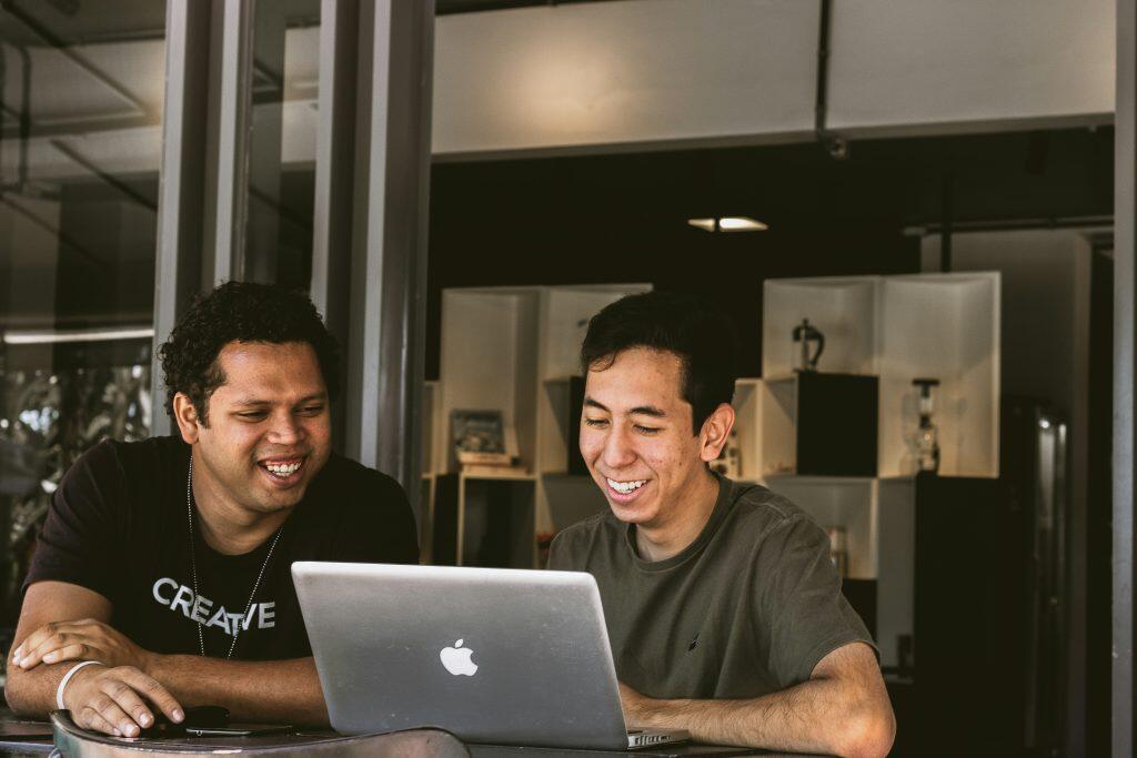 Two people sit side by side, smiling and looking at a silver Apple laptop in front of them. The person on the left wears a black t-shirt, while the person on the right wears a plain dark t-shirt. The background features shelves with books and decorative items, along with vertical beams and glass panels, suggesting a modern and collaborative workspace. Fiverr digital marketing.