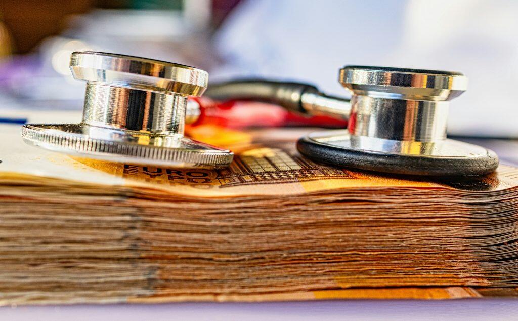 A stethoscope with a shiny metal chest piece and black rubber tubing rests on a stack of orange Euro banknotes. The warm lighting highlights the reflective surfaces of the stethoscope, emphasizing the focus on health insurance for small business, healthcare costs, or economic health.