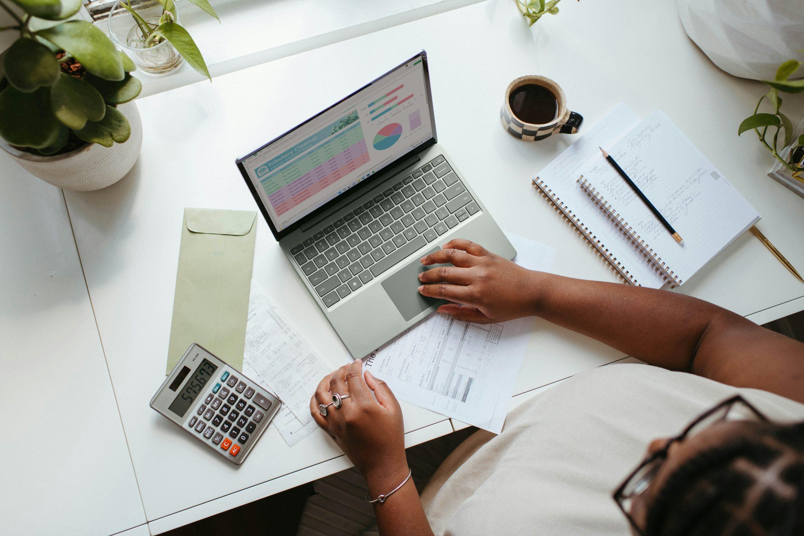 A clean, organized workspace with a white desk featuring a laptop displaying a colorful spreadsheet with a pie chart and bar graph. A person uses the trackpad with their right hand. To the left is a calculator and to the right, an open notebook with handwritten notes and a pencil. A coffee cup sits near the notebook, and a green envelope is partially visible by the calculator. Plant pots in the corners add a professional, inviting touch—perfect for financial or data analysis tasks and understanding what spreadsheets to keep for a small business.