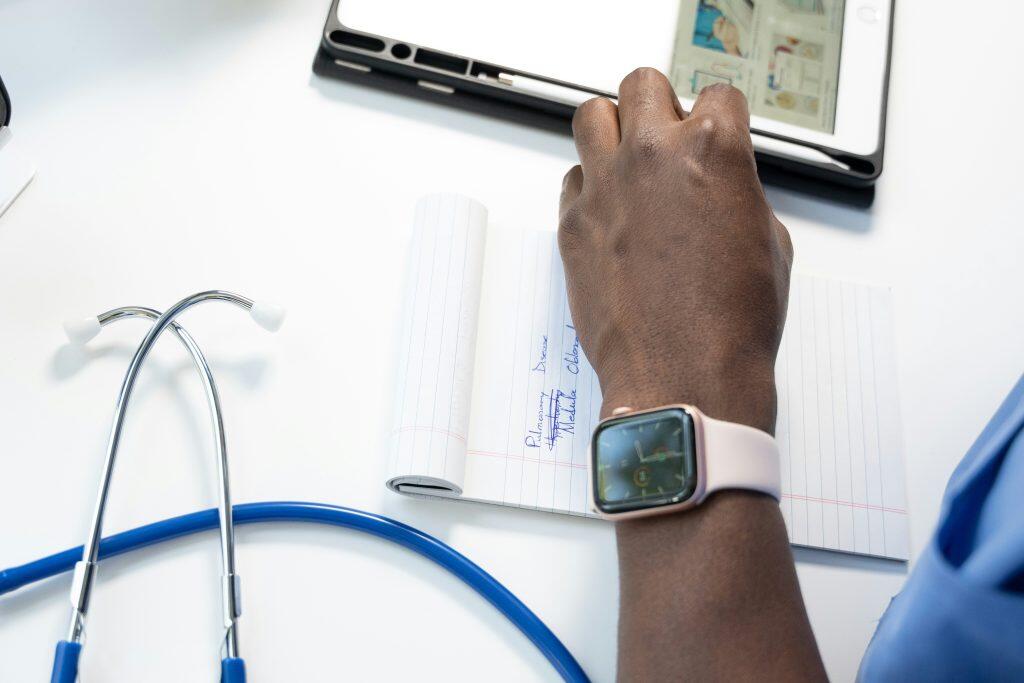 A medical workspace featuring a hand with a smartwatch writing on a notepad with handwritten notes about pulmonary disease and mediastinal oblong. A blue and silver stethoscope lies on the white surface, and a tablet with a partially visible screen is positioned above the notepad, suggesting a health insurance for small business or study setting.
