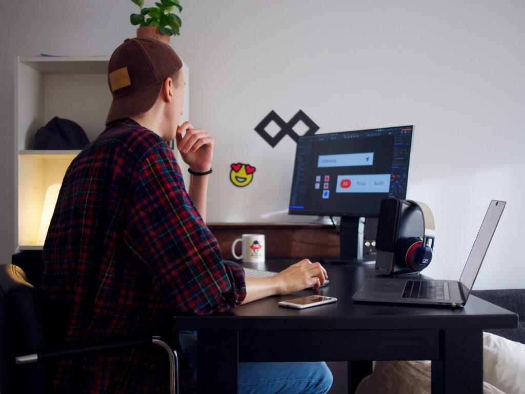 A person wearing a plaid shirt and a brown baseball cap sits at a dark wooden desk, working on a computer, perhaps weighing the pros and cons of Upwork vs Fiverr. The desk features a white mug with a red logo, a pair of headphones, a smartphone, a laptop, and a keyboard. The background includes a white wall with a geometric symbol and a smiley face sticker, as well as a shelving unit with a plant, a beanie, and a glowing light. The scene suggests a casual home office or creative workspace.