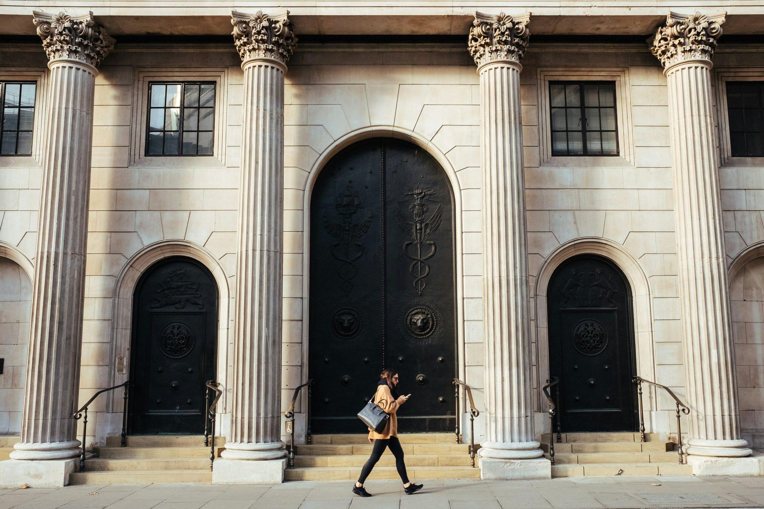 A classical architectural facade with five ornate Corinthian columns, three large black carved doors, and rectangular windows above each door. A person in a brown coat and black leggings walks in front of the central door, carrying a black bag and looking at their phone, perhaps researching the best banks for small business.