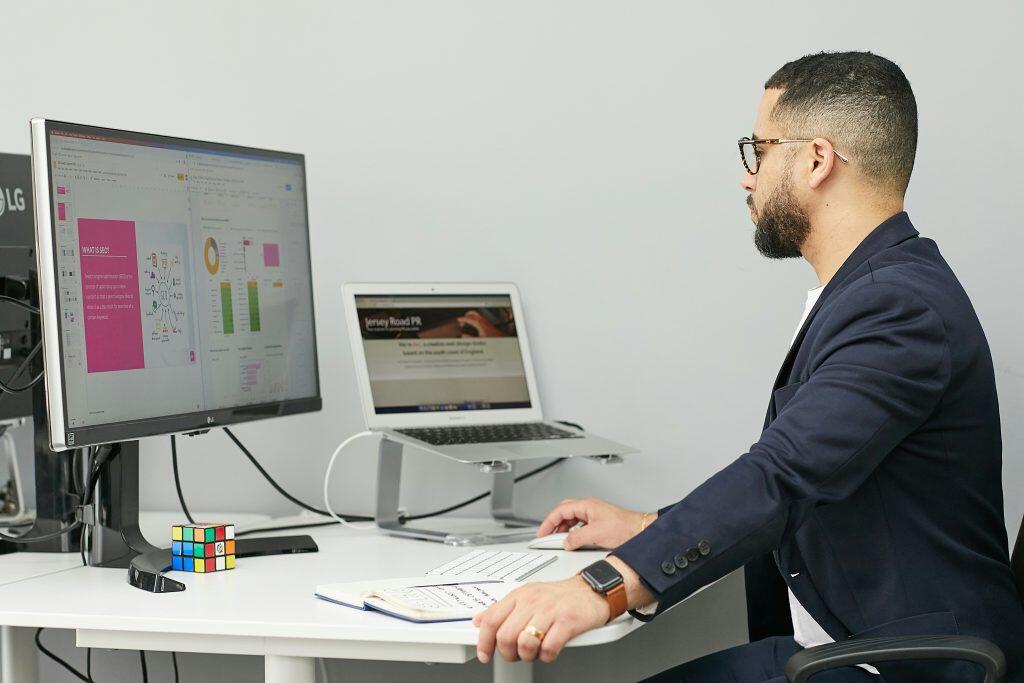 A workspace featuring a man with a beard and glasses seated at a desk. He’s wearing a dark blazer and smartwatch, working on a large monitor and laptop. The monitor shows an SEO presentation with a pie and bar chart, while the laptop displays a webpage. The desk is neatly arranged with a wireless keyboard, mouse, an open notebook with handwritten notes about what spreadsheets to keep for a small business, and a colorful Rubik's cube, adding a playful touch. The setting reflects a focused, tech-savvy work environment.