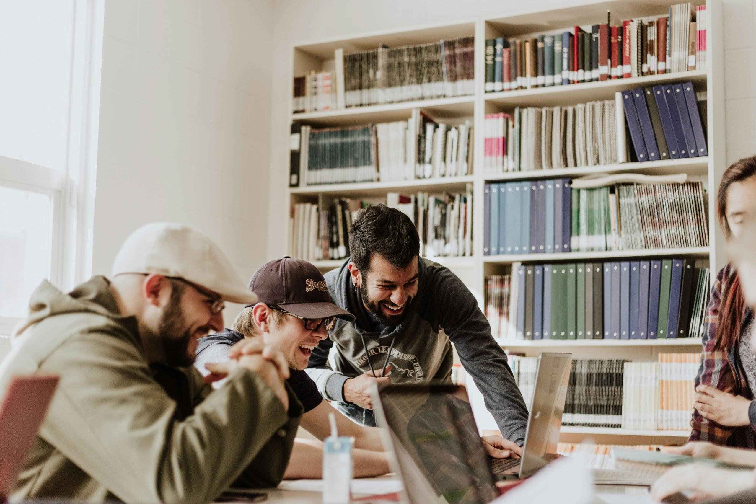 A group of four people collaborating around a table with laptops in a library-like setting, bookshelves filled with colorful binders in the background, conveying teamwork and a positive environment, relevant to Glassdoor reputation management.