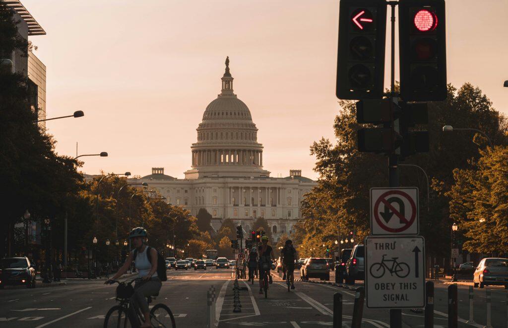 A serene urban scene in Washington, D.C., featuring the U.S. Capitol Building at sunset. The Capitol's iconic dome is centered in the background, framed by a warm orange-pink sky. A multi-lane road with a dedicated bike lane leads towards the Capitol, with bicyclists and cars in motion. Prominent traffic lights and signs, including a 'No U-Turn' and 'Obey This Signal' sign for bicyclists, are visible in the foreground. Lush green trees line the road, illuminated by the golden glow of the setting sun, creating a picturesque and nostalgic atmosphere, perhaps after a successful day at one of the many networking events in DC.