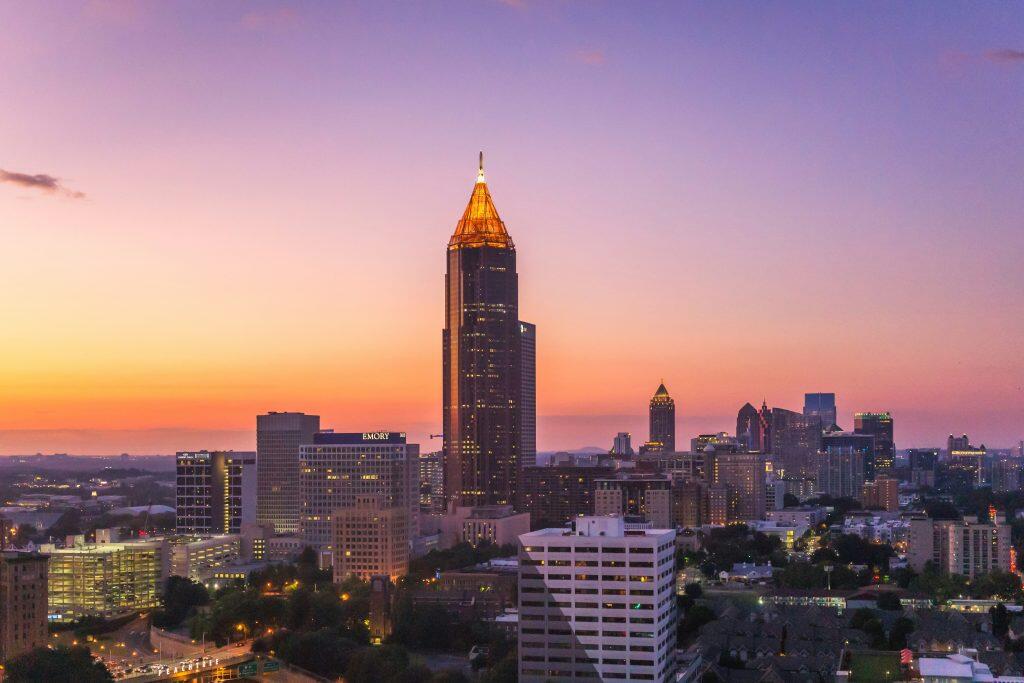 An aerial view of Atlanta's city skyline at sunrise or sunset, featuring a dramatic gradient sky transitioning from warm orange and pink near the horizon to deep purple at the top. The central focus is a tall skyscraper with a golden illuminated spire, surrounded by a dense array of buildings with glowing windows. Roads with streaks of car lights and patches of greenery are visible in the foreground, along with a partially visible green road sign reading 'PEACHTREE.' The vibrant cityscape also reflects the bustling energy of networking events Atlanta, connecting professionals amidst this dynamic urban hub.