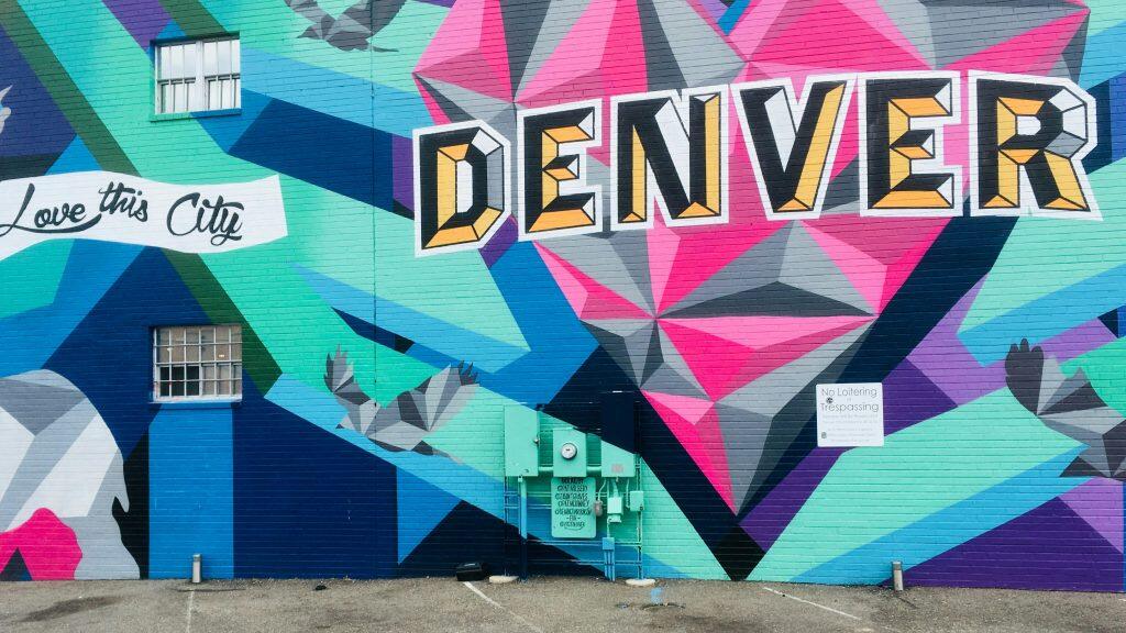 A vibrant mural on a brick wall in Denver, perfect for networking events Denver photography, featuring the phrase 'Love this City' in cursive on a white banner and the word 'DENVER' in large, three-dimensional block letters with orange and yellow shading. The mural showcases geometric patterns in cool tones of blue, green, and purple, accented with pink and black, along with stylized, angular birds. Below the mural, a white sign credits the artists and mentions '@VISITDENVER.'