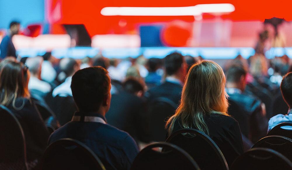 Trade show attendees watch a speaker on a convention center stage