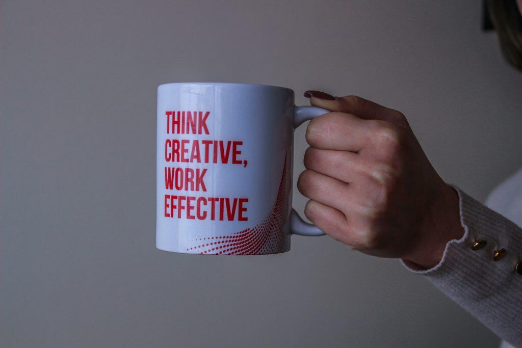 A white mug with bold red text reading 'THINK CREATIVE, WORK EFFECTIVE,' held by a hand wearing a light sweater with gold buttons, against a neutral gray background, highlighting the importance of choosing the right marketing materials photos vs videos static vs dynamic for impactful branding.