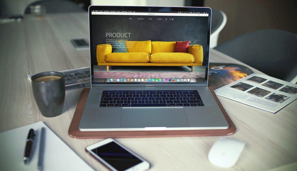 A modern workspace featuring an open silver MacBook Pro displaying a product-focused webpage with a vibrant yellow sofa and German text. The laptop rests on a brown leather mat, surrounded by a dark gray ceramic mug, a white notebook with pens, an open magazine, a white smartphone, and a computer mouse. The setup is on a light wooden table with visible grain, and the blurred background hints at chairs and office decor. The scene is well-lit, suggesting a clean and productive environment, reflecting design trends for 2025 in web design and graphic design.
