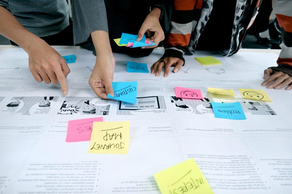 Overhead view of a collaborative UX design session at a desk. The workspace features a large paper diagram illustrating how to research if potential new product would sell, surrounded by colorful sticky notes with handwritten keywords like 'CUSTOMER,' 'Problem,' and 'JOURNEY MAP.' Multiple hands are visible, actively pointing, arranging sticky notes, and interacting with the diagram. The scene conveys teamwork and brainstorming, focusing on user experience design and problem-solving.