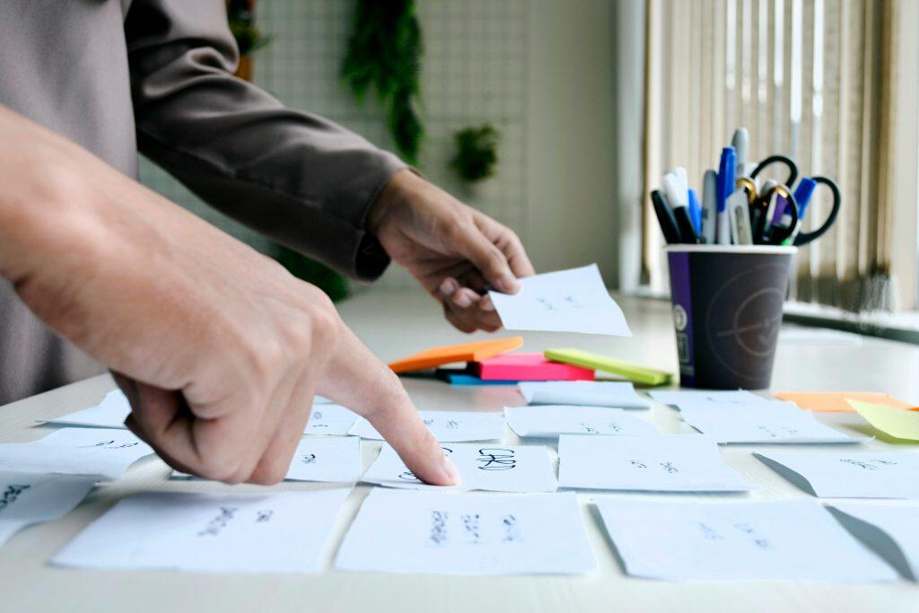 Close-up, high-angle view of hands organizing small white paper cards on a light-colored table. The left hand points to a card, while the right hand holds another card. The table features a dark cup filled with pens, markers, and scissors, as well as a stack of colorful sticky notes in orange, pink, and lime green. The background is blurred, showing hints of a plant and an indoor workspace, suggesting a brainstorming or organizational activity about how to research if potential new product would sell.