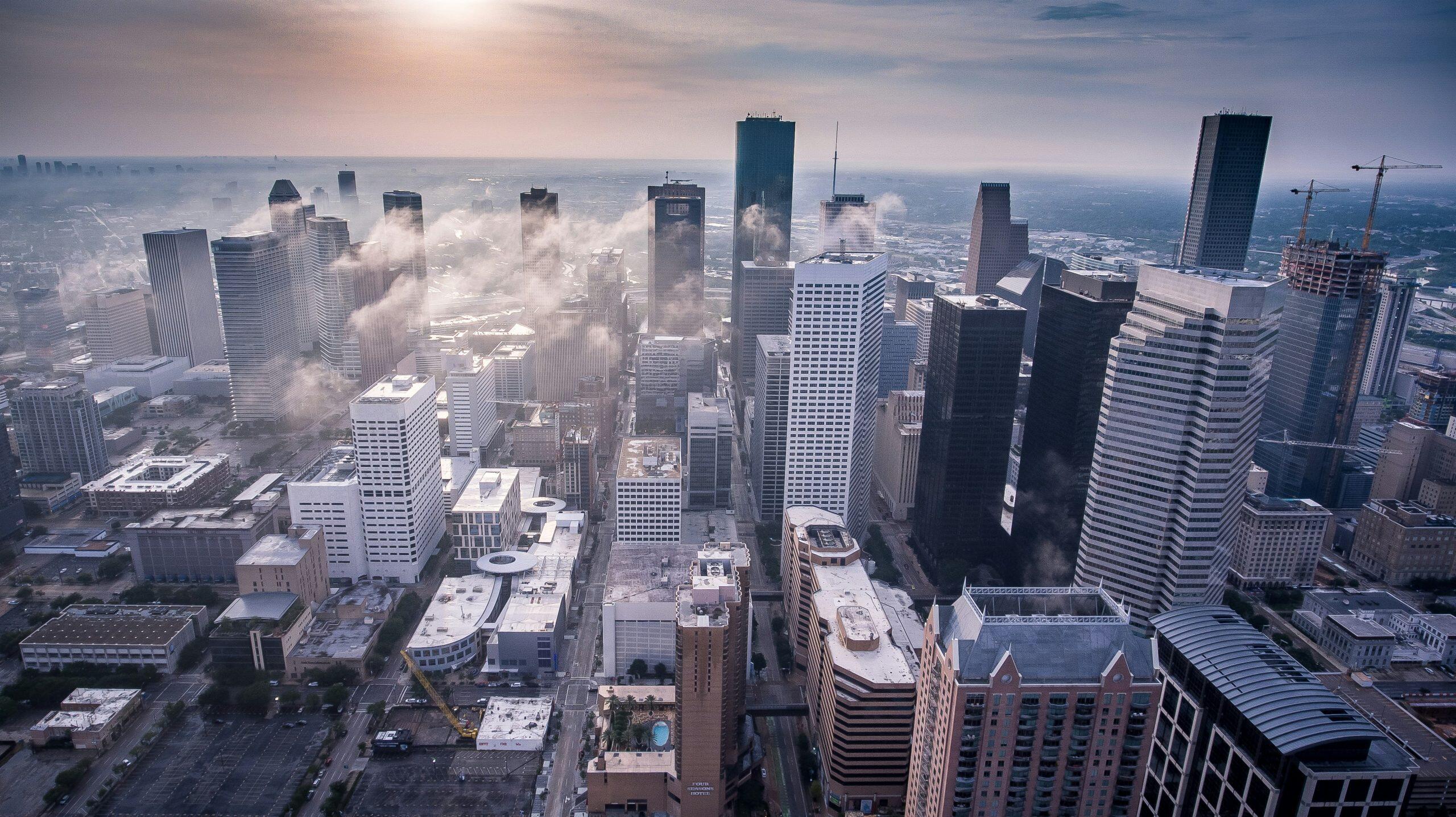 An aerial view of a bustling city skyline with numerous skyscrapers of varying heights and architectural styles. The scene features dense urban development, roads forming a grid-like layout, and cranes on the right side indicating ongoing construction. Fog or low clouds weave through the buildings, adding a dramatic atmosphere. The sky is overcast with a faint glow, suggesting early morning or late afternoon. A swimming pool is visible on a rooftop in the foreground, and the overall color palette is dominated by cool blues and greys, capturing the dynamic backdrop often seen during networking events Houston.
