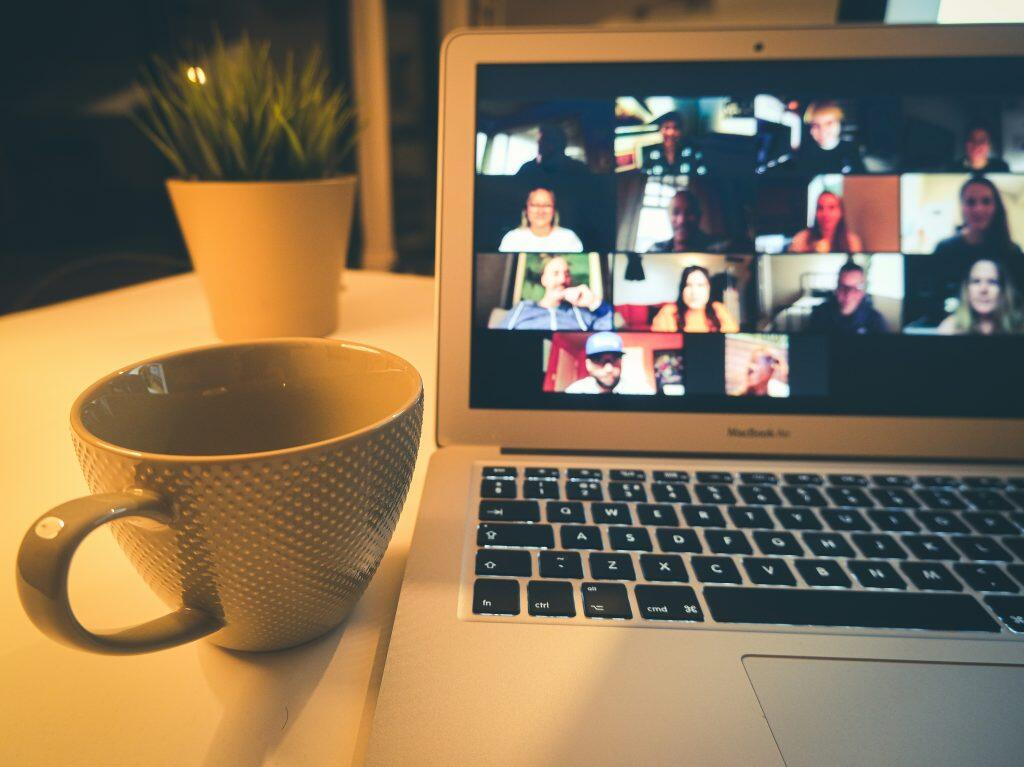 A cozy desk setup featuring a laptop displaying a video conference with multiple participants, a textured ceramic mug on the left, and a small potted plant in the background. The scene is illuminated by warm, ambient lighting, creating a comfortable and inviting atmosphere, often associated with remote work or virtual meetings—best way to keep up on social media trends.