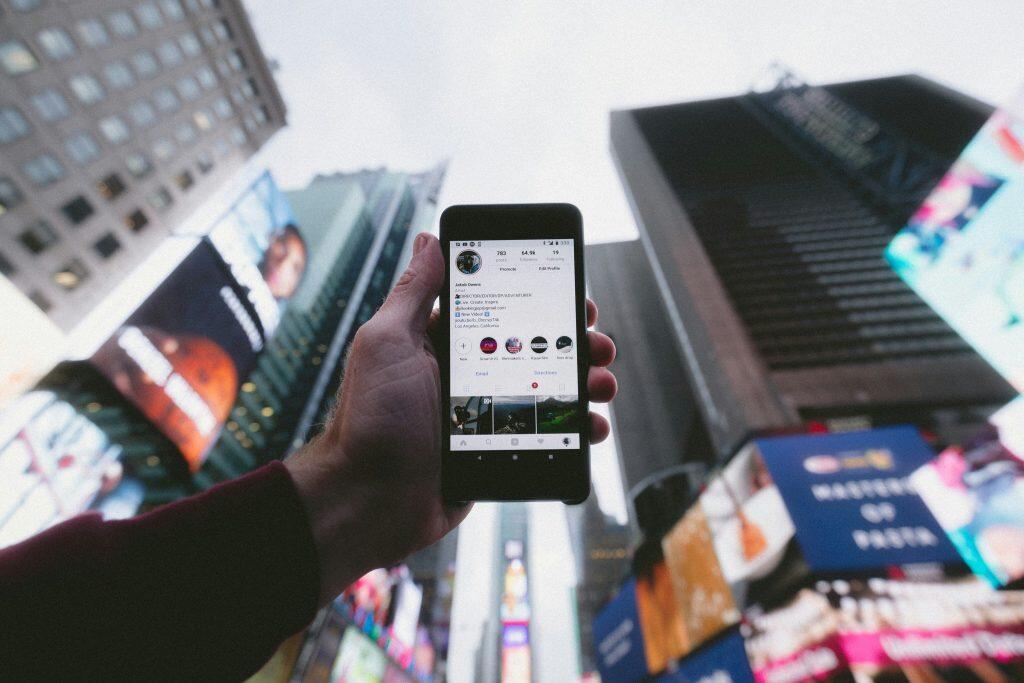 A close-up of a hand holding a smartphone displaying an Instagram profile against a vibrant urban backdrop of illuminated billboards and tall buildings, suggesting the best way to keep up on social media trends in a bustling, modern environment.