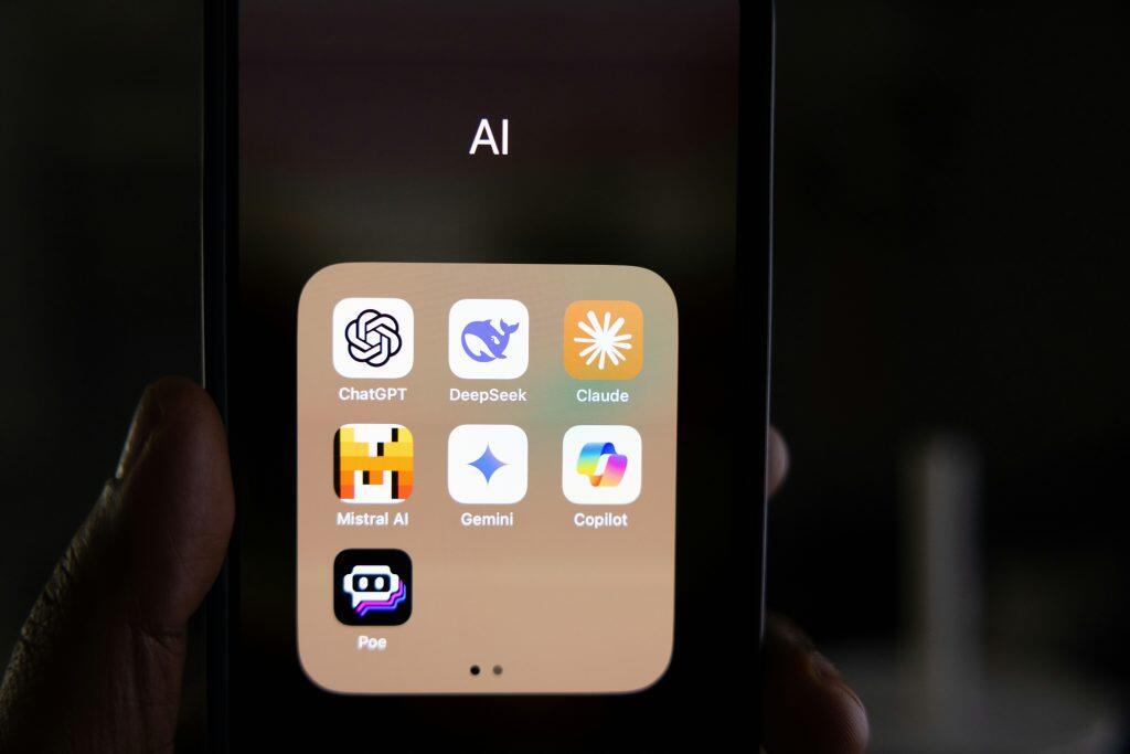 A close-up of a smartphone screen displaying an app folder titled 'AI,' containing seven app icons arranged in a grid. The apps include ChatGPT, DeepSeek, Claude, Mistral AI, Gemini, Copilot, and Poe, each with distinct logos and names. A dark-skinned hand holds the phone on the left side, with a blurred, dark background emphasizing the screen's content. The image highlights the organization of AI tools on a personal device, proving that smart organization is truly the best way to keep up on social media trends.