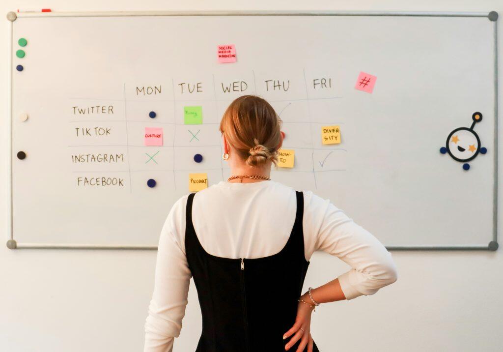 A person stands in front of a large whiteboard organized as a social media content calendar, a perfect example of the best way to keep up on social media trends. The board features a grid with days of the week as columns and social media platforms (Twitter, TikTok, Instagram, Facebook) as rows. Colored sticky notes with labels like 'Funny,' 'Culture,' and 'Product' mark planned content, while checkmarks, X's, and dots indicate task statuses. The person, wearing a black sleeveless top over a white shirt, observes the board with their hand on their hip. The scene conveys a structured and creative approach to social media marketing planning.