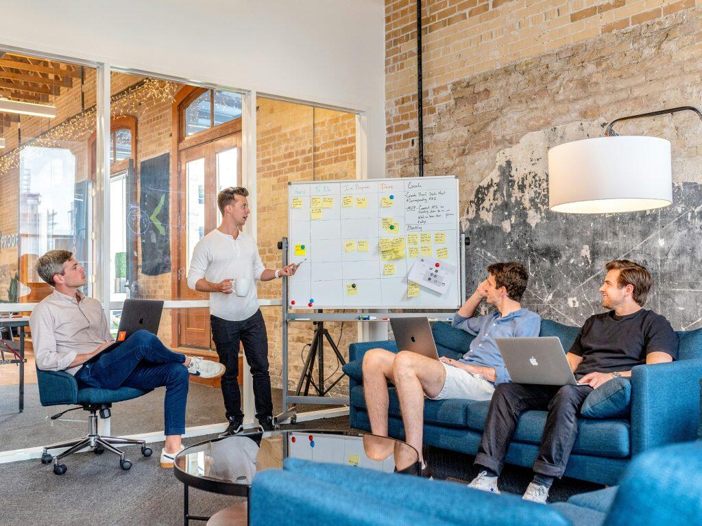 A collaborative office meeting with four individuals. One person stands near a whiteboard, presenting or explaining, while three others sit on a blue couch and chair, two using laptops. The whiteboard features sticky notes, handwritten notes, and sections labeled 'Story,' 'To Do,' 'In Progress,' and 'Done,' along with a 'Goals' section listing tasks like 'Complete Project Plan' and 'Meet with Client.' The setting includes a glass wall with wooden frames, a large white lamp, and a reflective coffee table, creating a modern and casual workspace focused on teamwork and project planning for a content marketing agency.