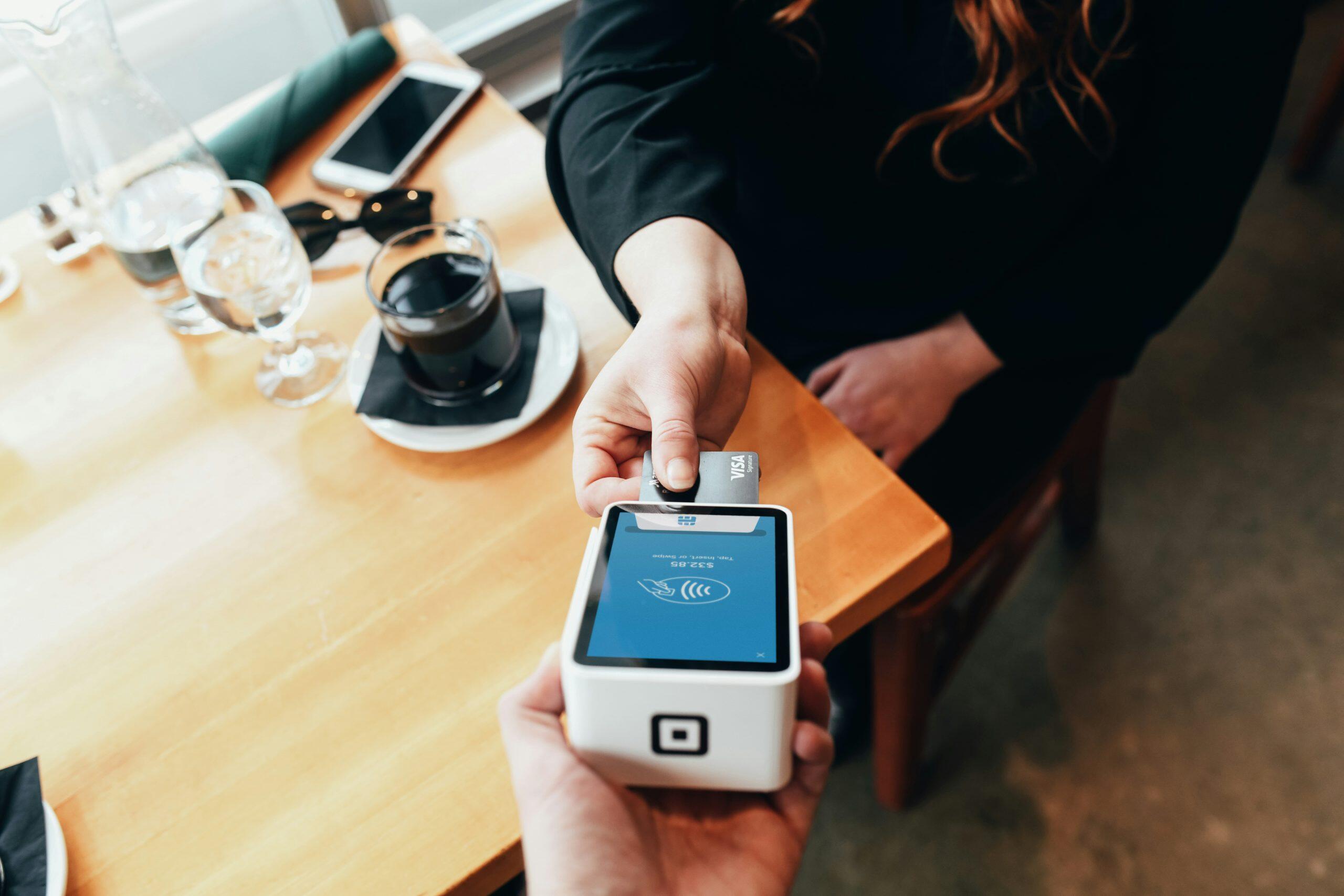A customer hands a Visa credit card to another person holding a small white card reader displaying a contactless payment symbol and a charge of $23.85. The transaction takes place on a wooden table in a café setting, with a cup of coffee, a glass of water, a smartphone, and sunglasses visible on the table. The scene highlights a modern, convenient payment experience, showcasing the best credit card processing for small business.