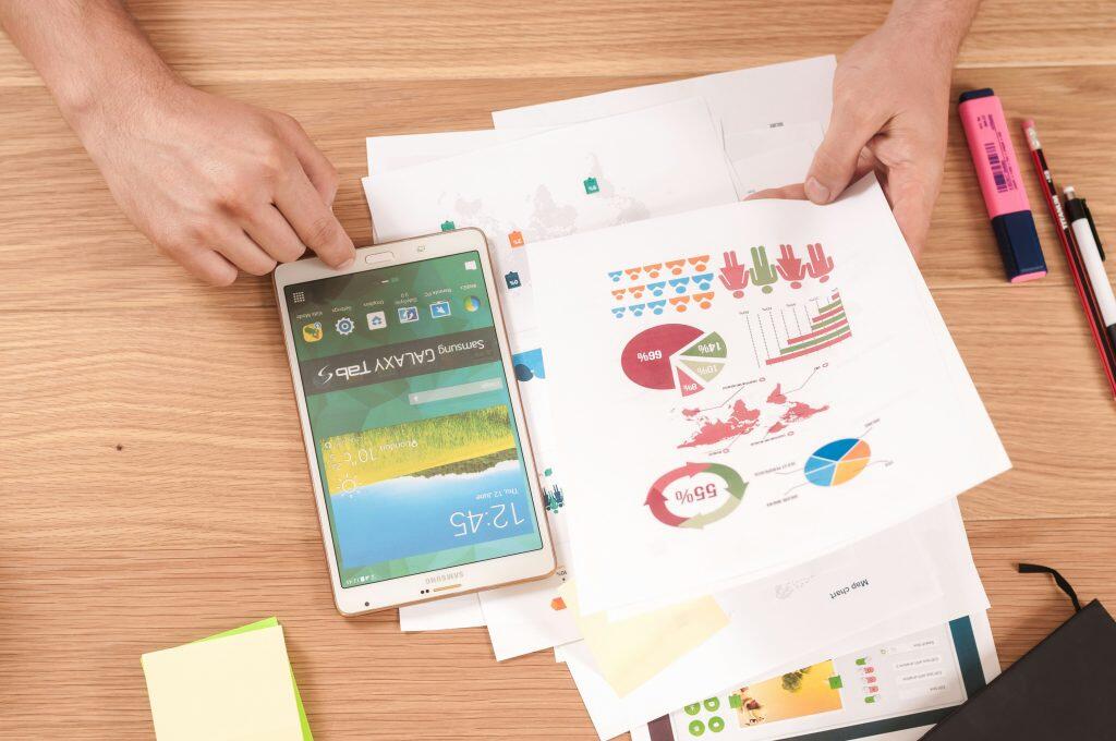 A workspace setup featuring a Samsung Galaxy Tab S placed upside down on a wooden desk. The tablet screen displays a home screen with app icons, a clock showing '12:45,' and the date 'Wednesday, July 16.' Surrounding the tablet are printed papers with colorful charts and graphs, including a pie chart, bar chart, and circular graph with percentages like '69%' and '60%.' Stationery items, such as a red pencil, black pencil with an eraser, pink highlighter, blue pen, and a stack of sticky notes, are scattered around. The scene suggests a focus on data analysis, report preparation, or content marketing agency business planning.