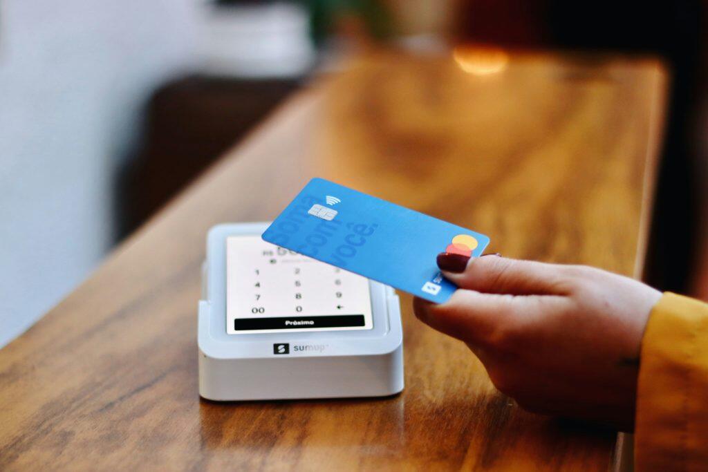 A blue credit card with a chip and contactless payment symbol is held by a hand, angled toward a white SumUp payment terminal. The terminal displays a numeric keypad and a button labeled 'Próximo,' sitting on a wooden countertop. The scene emphasizes a modern, user-friendly contactless payment experience, showcasing the best credit card processing for small business.