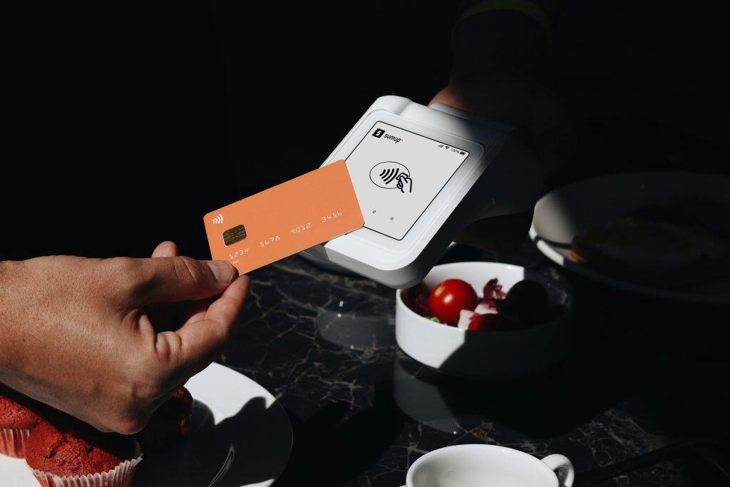 An orange credit card with a chip and contactless payment symbol is held near a white SumUp payment terminal displaying a contactless payment icon on its screen. The transaction takes place in a dining setting, with a plate of red muffins, a bowl of salad with cherry tomatoes, and a cup visible around the scene. The image highlights the ease and modernity of contactless payments in a café or restaurant environment, showcasing the best credit card processing for small business.