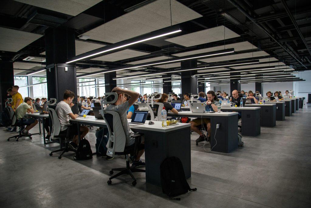 A modern open-plan office with rows of white desks, ergonomic chairs, and people working on laptops, illuminated by LED ceiling lights, with a casual and collaborative atmosphere, where discussions about exit interview questions occasionally take place.
