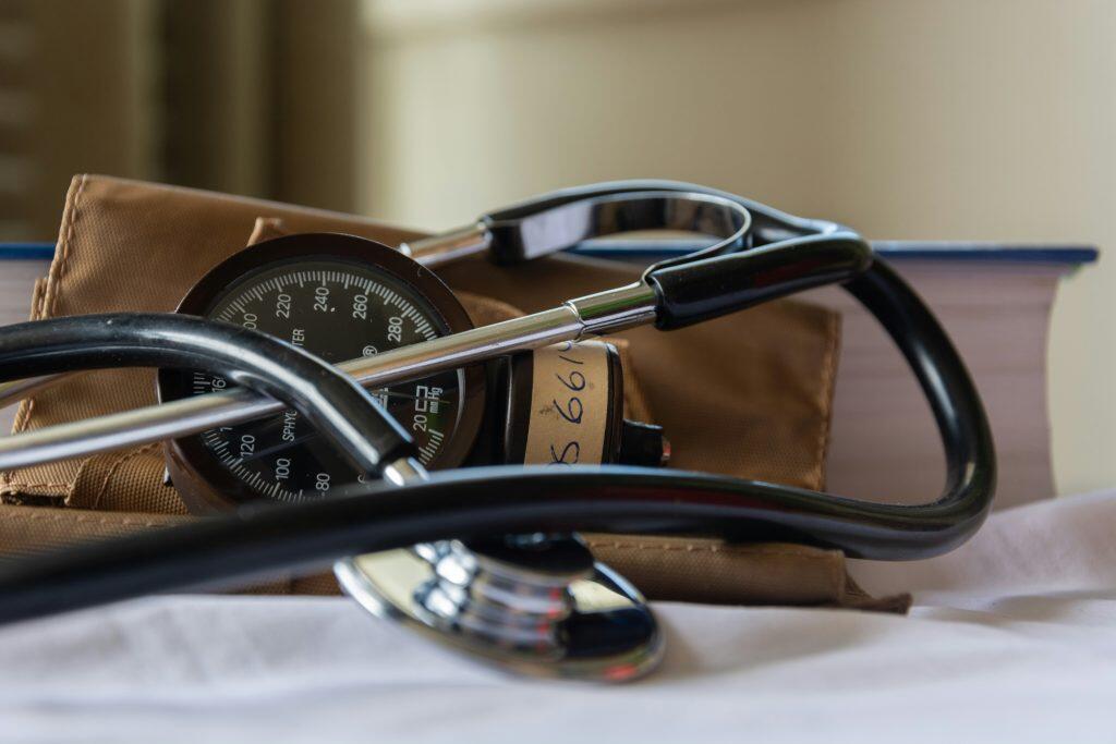 A close-up of medical instruments including a sphygmomanometer with a circular dial labeled 'mmHg' and a stethoscope with black tubing and a metallic chest piece. A brown blood pressure cuff is partially visible beneath the dial. A blue hardback book is blurred in the background, suggesting a medical or educational setting. This highlights the importance of health insurance for small business in ensuring access to essential tools and resources.