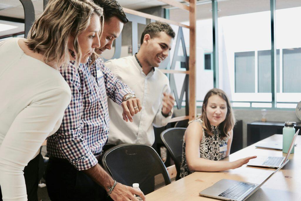A group of five colleagues from an employee benefits corporation collaborates around a wooden table in a bright, modern office. Three individuals are standing and leaning over to view a laptop screen, while two others are seated and engaged with their own laptops. The scene suggests a productive team meeting or brainstorming session, with a relaxed and positive atmosphere. The background features large windows, a wooden shelving unit, and modern decor elements.