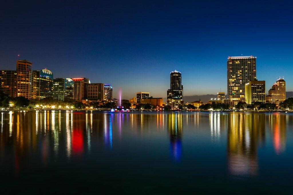 A vibrant nighttime city skyline reflected in a calm body of water. The scene features illuminated buildings with warm yellow, orange, blue, and red lights, a colorful fountain in the center, and shimmering reflections on the water. The dark blue sky contrasts with the bright city lights, creating a tranquil and picturesque urban landscape—perfect for networking events Orlando.