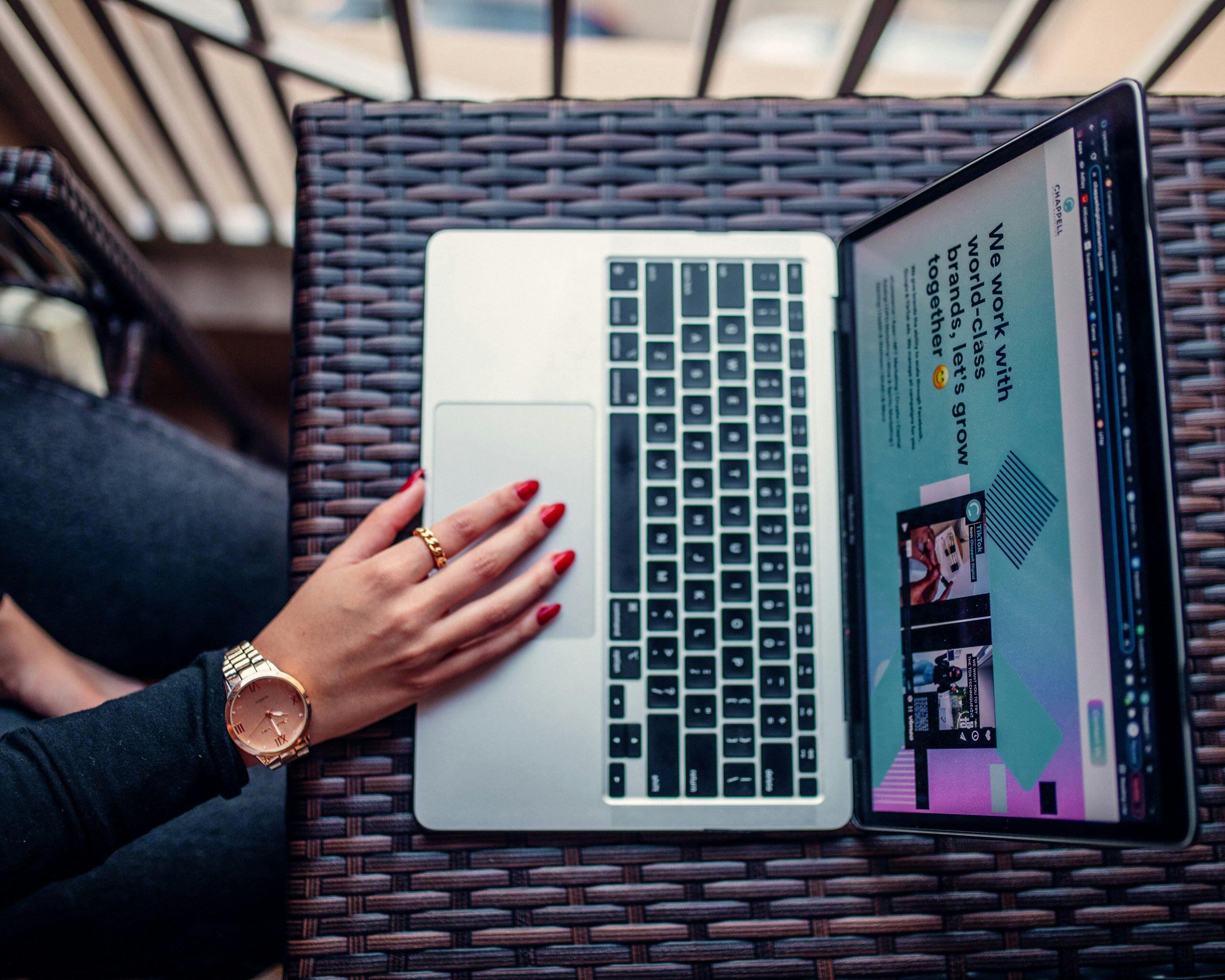 A stylish hand with red-painted nails, wearing a gold watch and rings, rests on a laptop trackpad on a dark wicker table, with the laptop screen displaying a webpage about brand collaboration and the best website builder for small business.