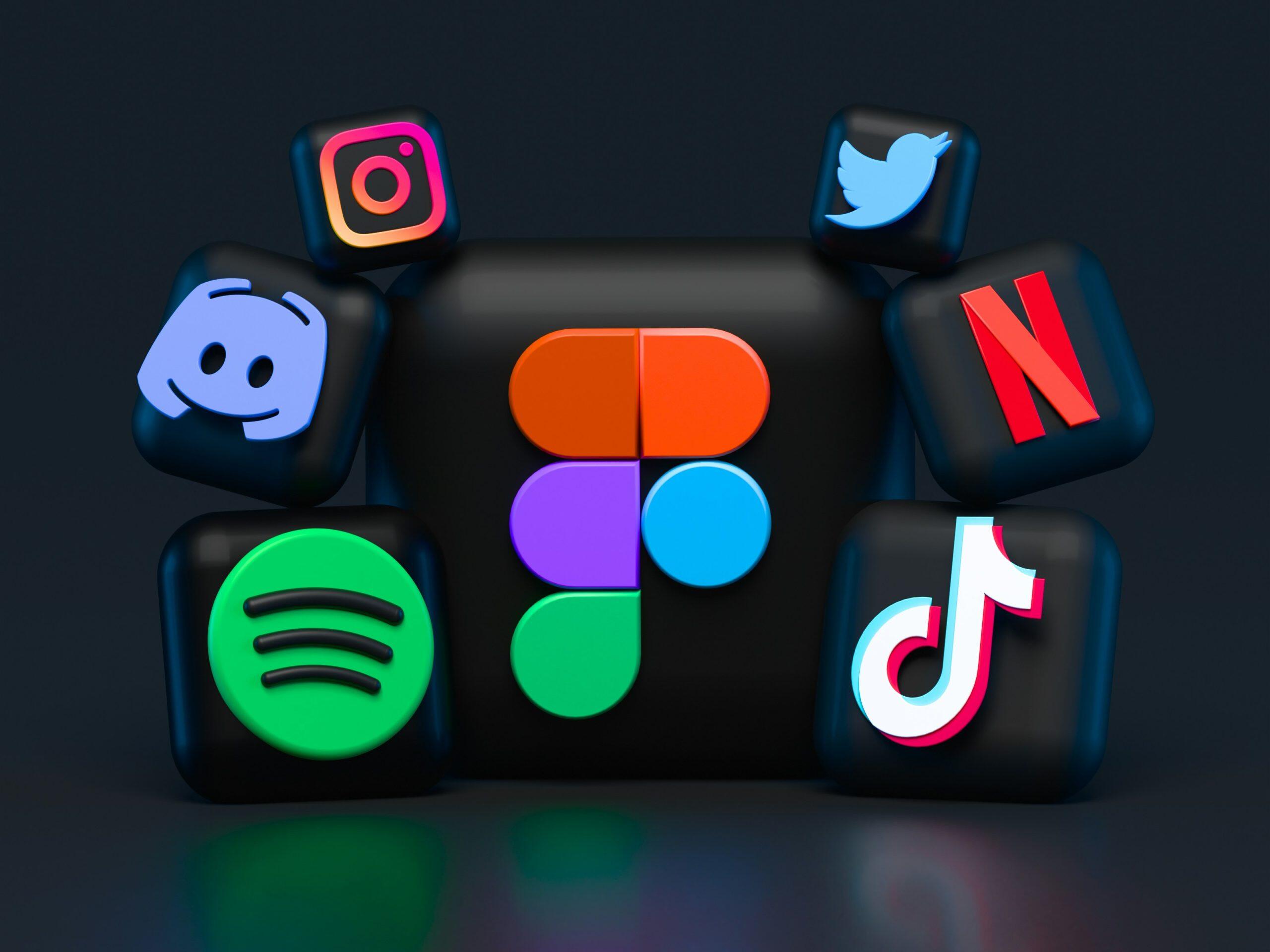 A 3D-rendered image featuring a central matte black block with the colorful Figma logo, surrounded by six smaller black blocks displaying logos of popular apps: Discord, Instagram, Twitter, Netflix, TikTok, and Spotify, which can drive social media reviews. The blocks rest on a reflective dark surface, with subtle color reflections from the logos. The background is a solid dark grey/black, emphasizing the vibrant colors of the logos.