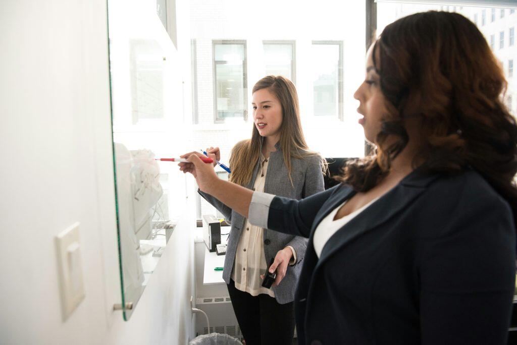 Two women collaborate in a modern office setting with a large glass whiteboard and a bright window in the background. The Client Success Manager in the foreground, with long wavy dark brown hair, wears a navy blazer over a white top and holds a red marker, writing or drawing on the whiteboard. The woman in the background, with straight brown hair, wears a light grey blazer over a cream button-up shirt and holds a blue marker, observing the activity. A white desk beneath the whiteboard holds black electronic devices and cables, while a large window floods the room with natural light, revealing the exterior of a modern building.