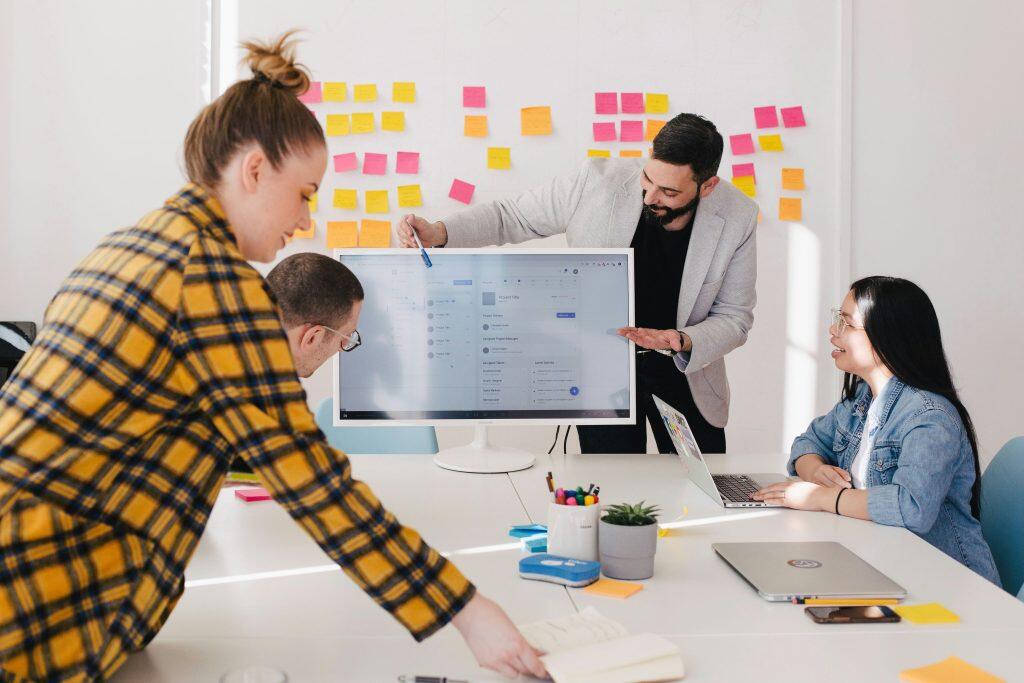 A collaborative office setting with four individuals engaged in a team discussion. A person in a light-colored blazer stands and points at a large monitor displaying a project management interface, a tool often used in direct response marketing, while others observe or work on laptops. The white table is scattered with office supplies, including a small potted plant, colorful pens, a notebook, and sticky notes. In the background, colorful post-it notes are affixed to the wall, suggesting brainstorming or planning activities often seen in direct response marketing strategies. The scene is well-lit, emphasizing a professional and interactive work environment.