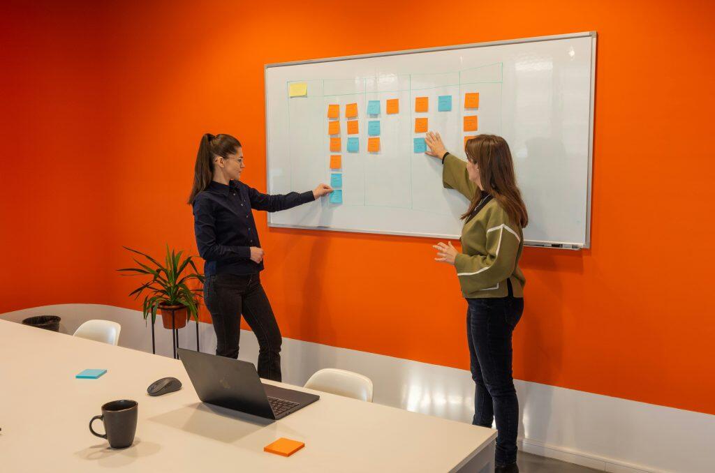 Two women collaborate in a modern office space with a vibrant orange wall and a large whiteboard covered in color-coded sticky notes. The woman on the left, with dark brown hair in a ponytail, wears glasses and a dark blue collared shirt, pointing to a blue sticky note on the whiteboard. The Client Success Manager on the right, with long brown hair, wears an olive green sweater with striped sleeves, gesturing near an orange sticky note. The whiteboard is divided into a grid, resembling a project management board, with notes. A table in the foreground holds a laptop, sticky note pads, a mug, and a potted plant, adding to the collaborative and organized atmosphere.