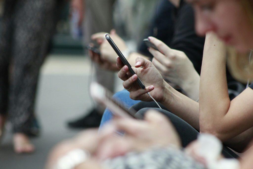 A close-up shot of multiple hands holding smartphones, with a central focus on a hand holding a dark smartphone connected to a white charging cable. The background and surrounding elements, including other hands, devices, and blurred figures, suggest a group of people engaged with their phones in a casual indoor setting. The image emphasizes digital connectivity and modern technology usage to drive social media reviews.