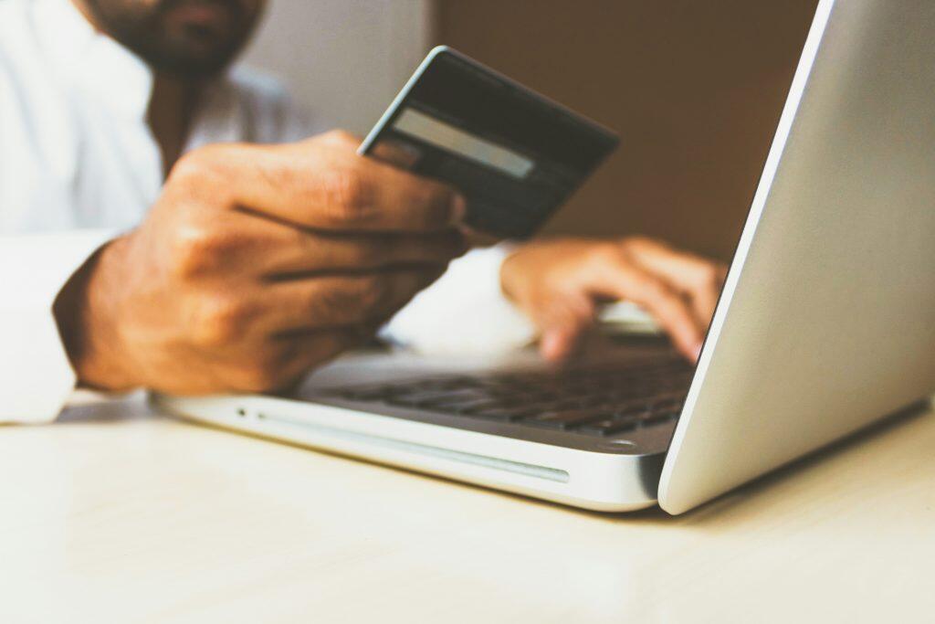 A close-up shot of a person holding a dark credit card in their right hand above a silver laptop on a light-colored desk. The laptop's keyboard and screen are slightly out of focus, suggesting active use. The background is blurred, showing warm tones and the faint outline of the person, emphasizing the act of online payment or drive social media reviews or digital transaction.