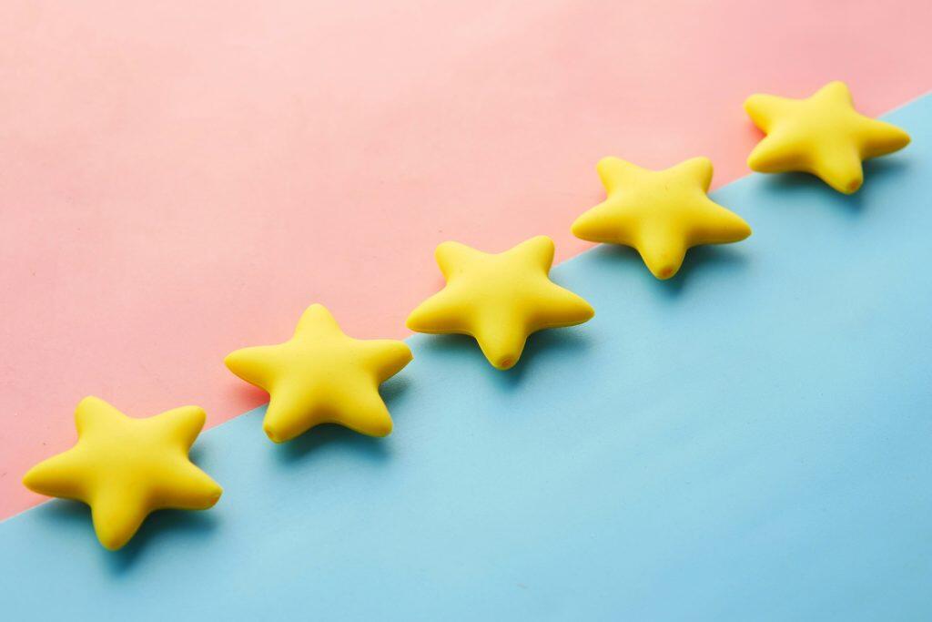 Six yellow, plump, five-pointed stars arranged diagonally across a background split into two pastel colors: light pink in the upper-left and light blue in the lower-right. The stars have a soft, rounded appearance with subtle shadows, adding depth. The diagonal alignment of the stars mirrors the sharp division of the background colors, creating a harmonious and cheerful composition, designed to drive social media reviews.