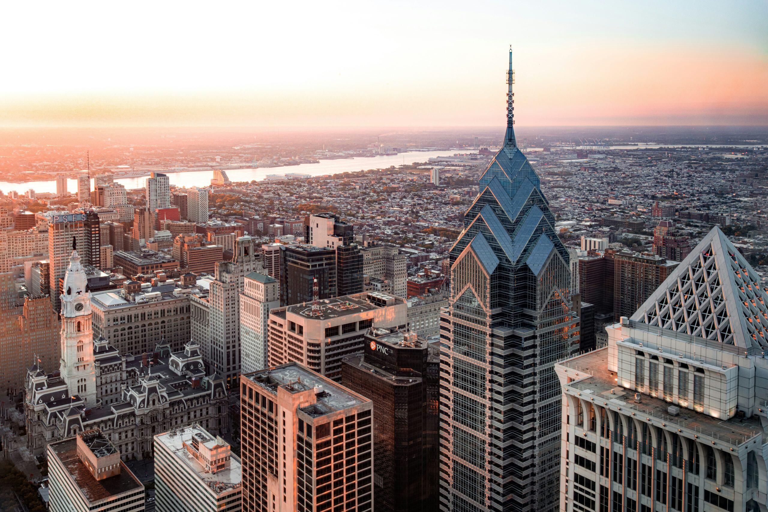 An aerial view of Philadelphia at sunrise or sunset, showcasing a sprawling cityscape with a warm golden glow. Prominent landmarks include the Philadelphia City Hall with its clock tower and statue, and modern skyscrapers like One Liberty Place with its blue-tinted glass facade and spire. The city is layered with dense urban buildings, transitioning to residential areas and industrial complexes near a wide river in the background. The sky is hazy, blending from bright yellow to soft pink hues, adding depth and atmosphere to the scene. This vibrant cityscape sets the perfect backdrop for networking events Philadelphia, where connections and opportunities flourish.