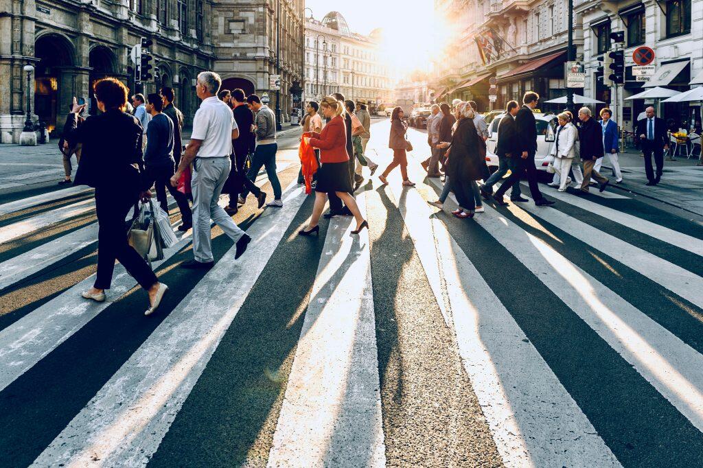 An urban street scene in Vienna during late afternoon or early evening, featuring a diverse group of pedestrians crossing a wide, striped crosswalk. The low sun casts long, dramatic shadows across the street, highlighting the bustling activity. Ornate historical buildings line the left side, while modern buildings with outdoor cafes and flags are on the right. A 'HOTEL' sign and 'Opernpassage' signage are visible, along with a few vehicles in the background. The warm sunlight creates a vibrant and dynamic atmosphere, reminiscent of the energy found at networking events Orlando.