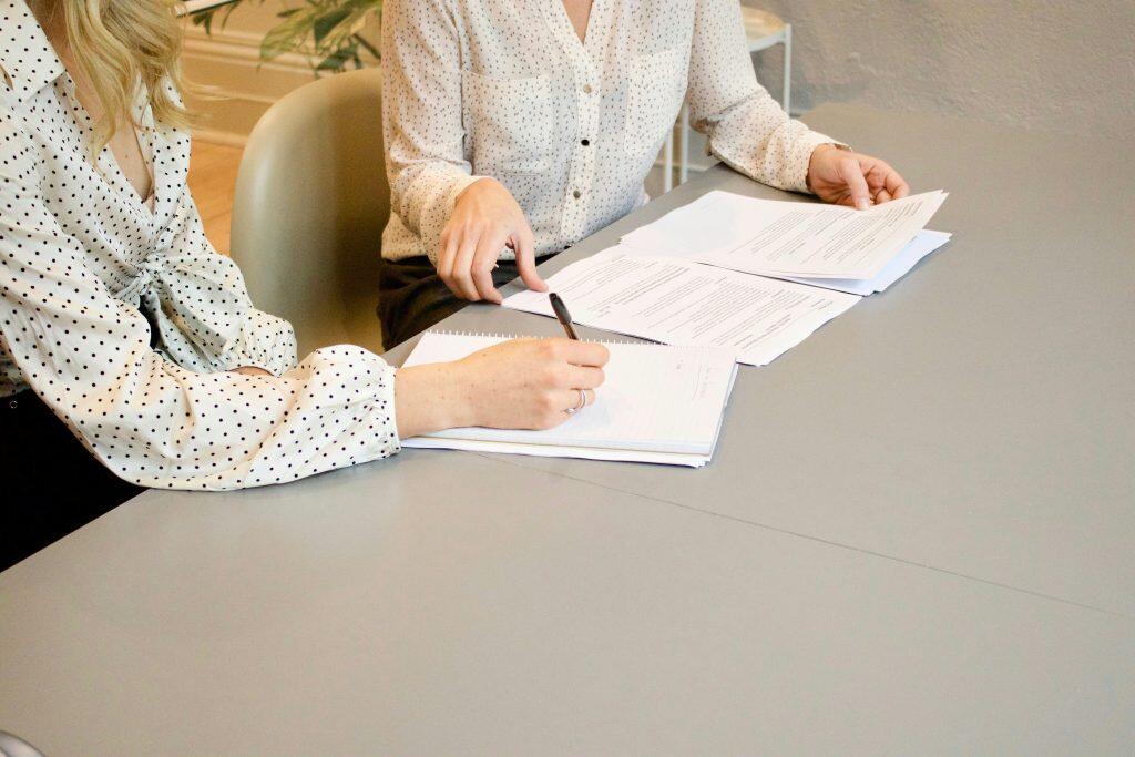 Two individuals seated at a grey table, engaged in a professional discussion. The person on the left, wearing a white polka dot shirt, is writing in a spiral-bound notebook with a pen. The person on the right, in a light-colored shirt with a subtle pattern, is reviewing a stack of documents. The table is covered with papers and a notebook, suggesting a collaborative task on topics like how to build business credit. The background is blurred, showing a light office chair and faint greenery, indicating an indoor office setting.
