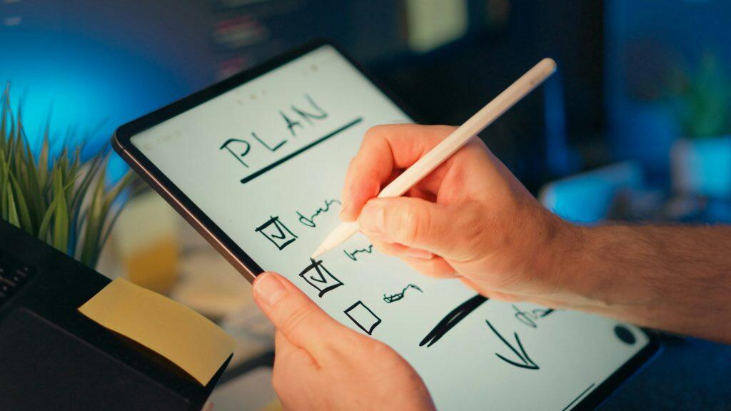 Close-up of a person's hands using a white digital stylus on a dark tablet screen. The screen displays a handwritten title 'PLAN' with three checkboxes below it, two of which are checked, and the third is empty. The right hand is actively marking a checkbox, while the left hand steadies the tablet. The blurred background features a green plant, a yellow sticky note, and a dark keyboard, with cool blue ambient lighting creating a modern workspace atmosphere. The image conveys themes of how to build business credit, digital planning, and productivity.