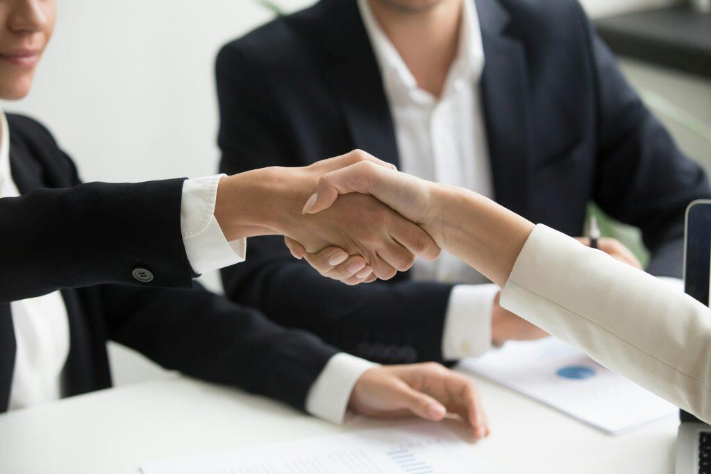 Close-up of a handshake between two individuals in a professional setting. The person on the left wears a black blazer over a white collared shirt, while the person on the right wears a light-colored blazer. A blurred background shows a third person in a dark suit seated at a white table with documents, including one featuring a blue pie chart, a pen, and a partially visible laptop. The image conveys themes of how to build business credit, agreement, partnership, and professionalism.