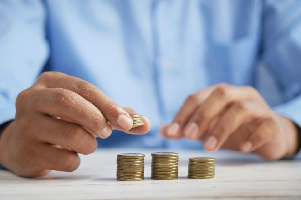 Close-up of a person's hands stacking golden coins on a light-colored surface. The left hand is holding a small stack of coins, poised to add to one of three neatly arranged coin stacks of varying heights. The background is blurred, showing a soft blue hue, likely from the person's shirt, emphasizing the coins and hands as the focal point. The image conveys themes of financial management, saving, and how to build business credit.