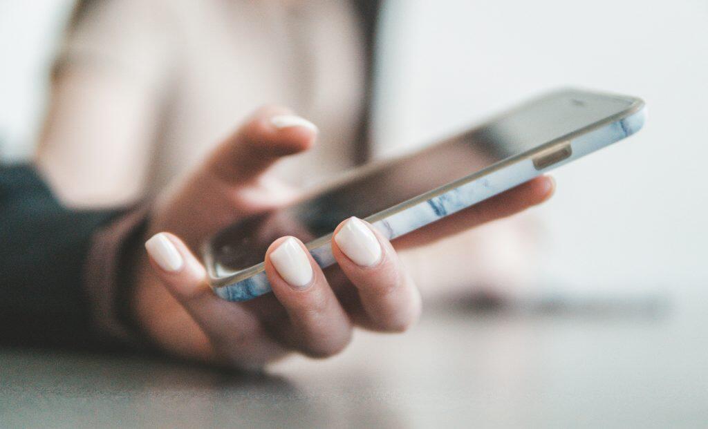 A close-up shot of a hand with manicured, light-colored nails holding a smartphone horizontally. The phone has a light blue and white marbled case, and its screen is reflective of what are voice search ads. The background is softly blurred, creating a bokeh effect, with muted tones and soft lighting highlighting the hand and phone.