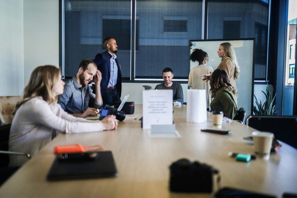 Casual office meeting with seven individuals in a collaborative workspace. Three people are seated at a large wooden conference table on the left, engaged in conversation or work, while two others stand near a whiteboard in the background. One person stands by the window, and another sits at the far end of the table, smiling. The table features a laptop, a camera, a stack of paper towels, and disposable coffee cups. Large windows with blinds provide natural light, and a potted plant adds greenery to the room. A sign on the table reads, 'Please clean dishes after use to maintain a clean meeting room. Thank you.' This setting could provide inspiration for SEO marketing for small business, showcasing a professional yet approachable environment.