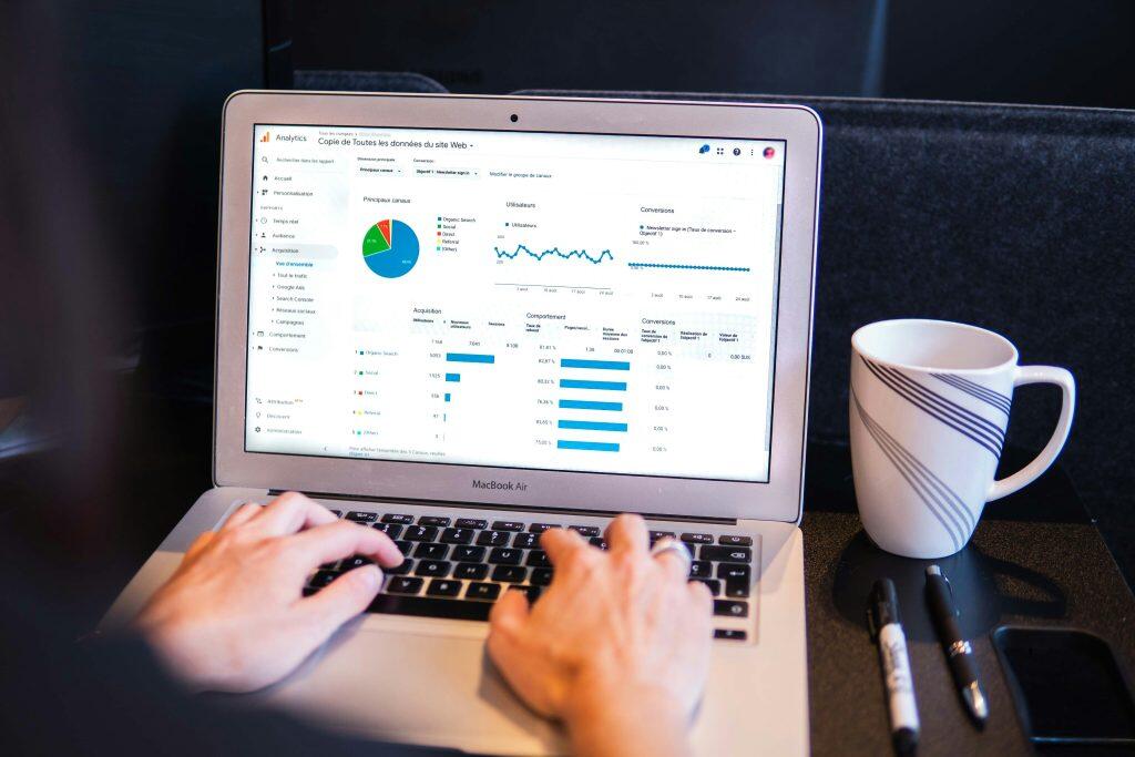 Person analyzing web traffic data on a MacBook Air displaying a web analytics dashboard, likely Google Analytics. The dashboard includes a pie chart showing traffic sources (Organic Search, Direct, Referral, Social, Other), a line graph illustrating user trends over time, and bar charts detailing acquisition, behavior, and conversion metrics. The environment features a white mug with blue diagonal stripes, two markers, and a smartphone on the table, suggesting a casual workspace—perfect for focusing on SEO marketing for small business strategies.