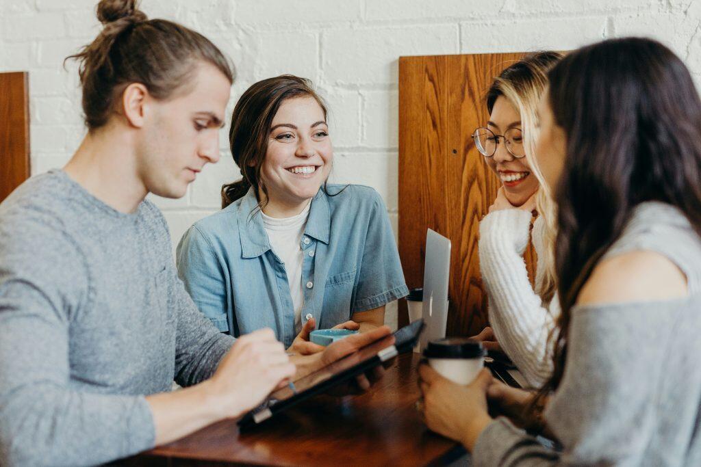 Four individuals sit around a table in a casual setting, likely a cafe, engaged in a lively conversation. One person holds a tablet, another has a laptop, and two hold coffee cups, suggesting a mix of work and social interaction. The background features wooden panels and a white brick wall, with warm lighting creating a friendly and collaborative atmosphere—perfect for networking events San Diego.