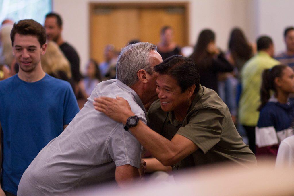 Two men warmly embrace in the foreground of an indoor setting, one wearing a light gray shirt and the other a dark green shirt, both smiling. A young man in a blue shirt looks on from the left, while several other individuals are visible in the softly blurred background, engaged in conversations. The scene conveys a friendly and welcoming atmosphere, likely at a community or social gathering, such as networking events San Diego.