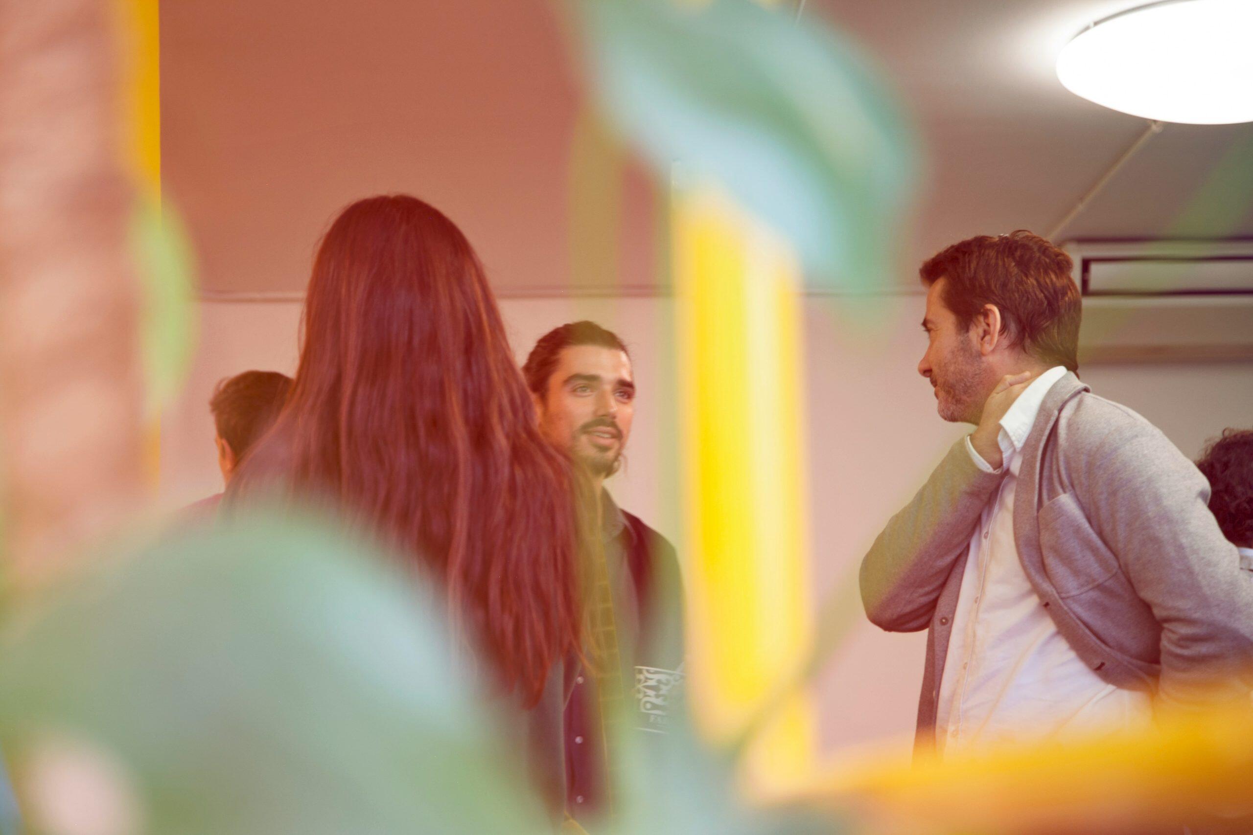 Three individuals are engaged in a casual conversation in an indoor setting with soft, warm lighting. The foreground features blurred green plants, and the background includes a circular ceiling light and air conditioning vents. The atmosphere is relaxed and informal, resembling a small meeting or networking events San Diego in an office or workspace.